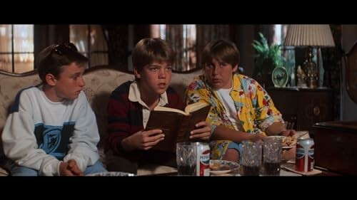 Three boys sitting on a sofa, one reading a book, with drinks on the table.