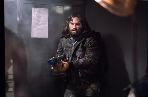 Man with long hair holding a gun in a dimly lit room.